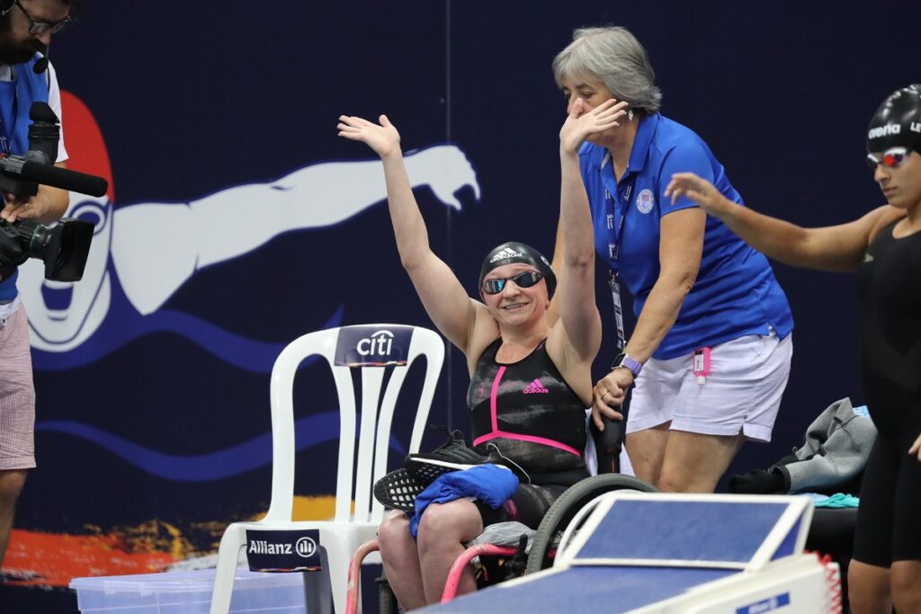 A female swimmer in a swimsuit and swim cap smiles and raises her arms while sitting in a wheelchair by the pool, assisted by a woman in blue. McKenzies Story shines as another swimmer and a cameraman appear nearby, with a mural of a swimmer in the background.