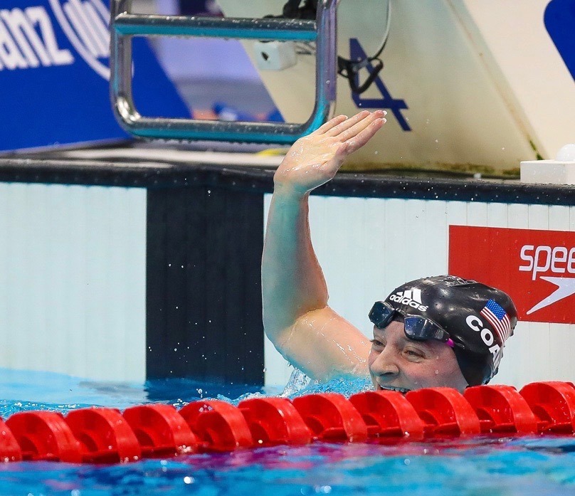 A swimmer wearing goggles and a swim cap smiles and waves while in the pool, next to the lane divider and end wall after finishing a race, capturing the spirit of McKenzies Story.