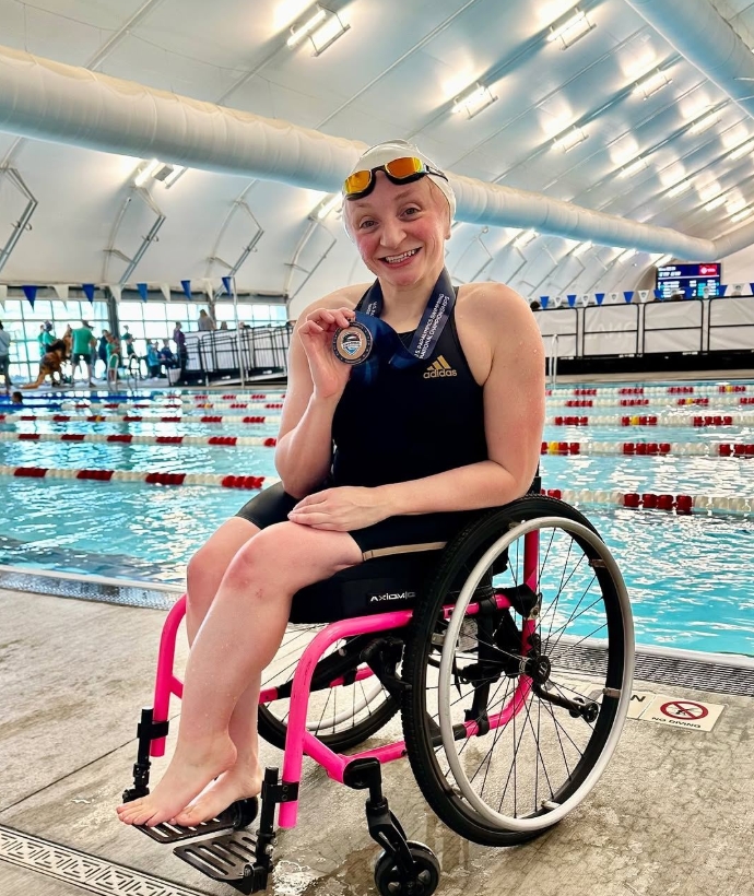 A smiling swimmer in a swimsuit and goggles sits in a pink wheelchair by an indoor pool, proudly holding up a medal with a ribbon. Swimming lanes and timing boards are visible in the background, celebrating McKenzies Story.
