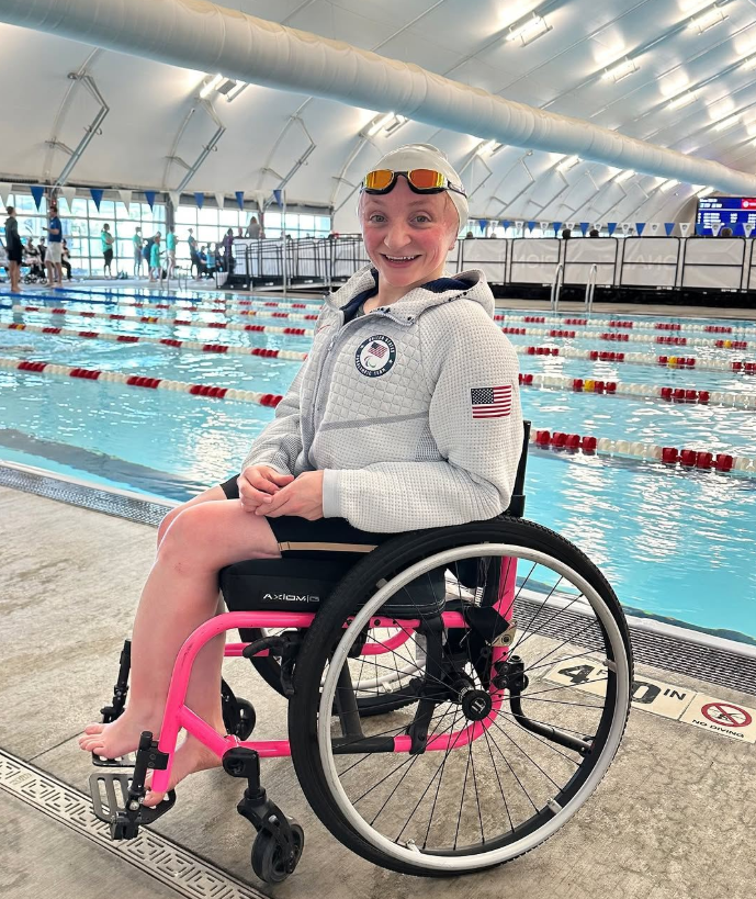 A smiling swimmer wearing goggles and a Team USA jacket sits in a pink wheelchair by an indoor swimming pool, with lanes and lane markers visible in the background—capturing the spirit of McKenzies Story.