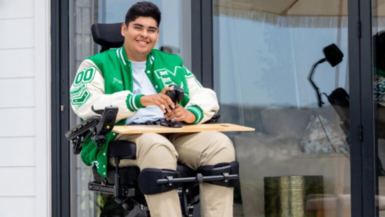 A young man in a motorized wheelchair, wearing a green and white varsity jacket, smiles while holding a game controller. He benefits from custom contoured seating, and is seated outside near sliding glass doors with a tray attached to his chair.