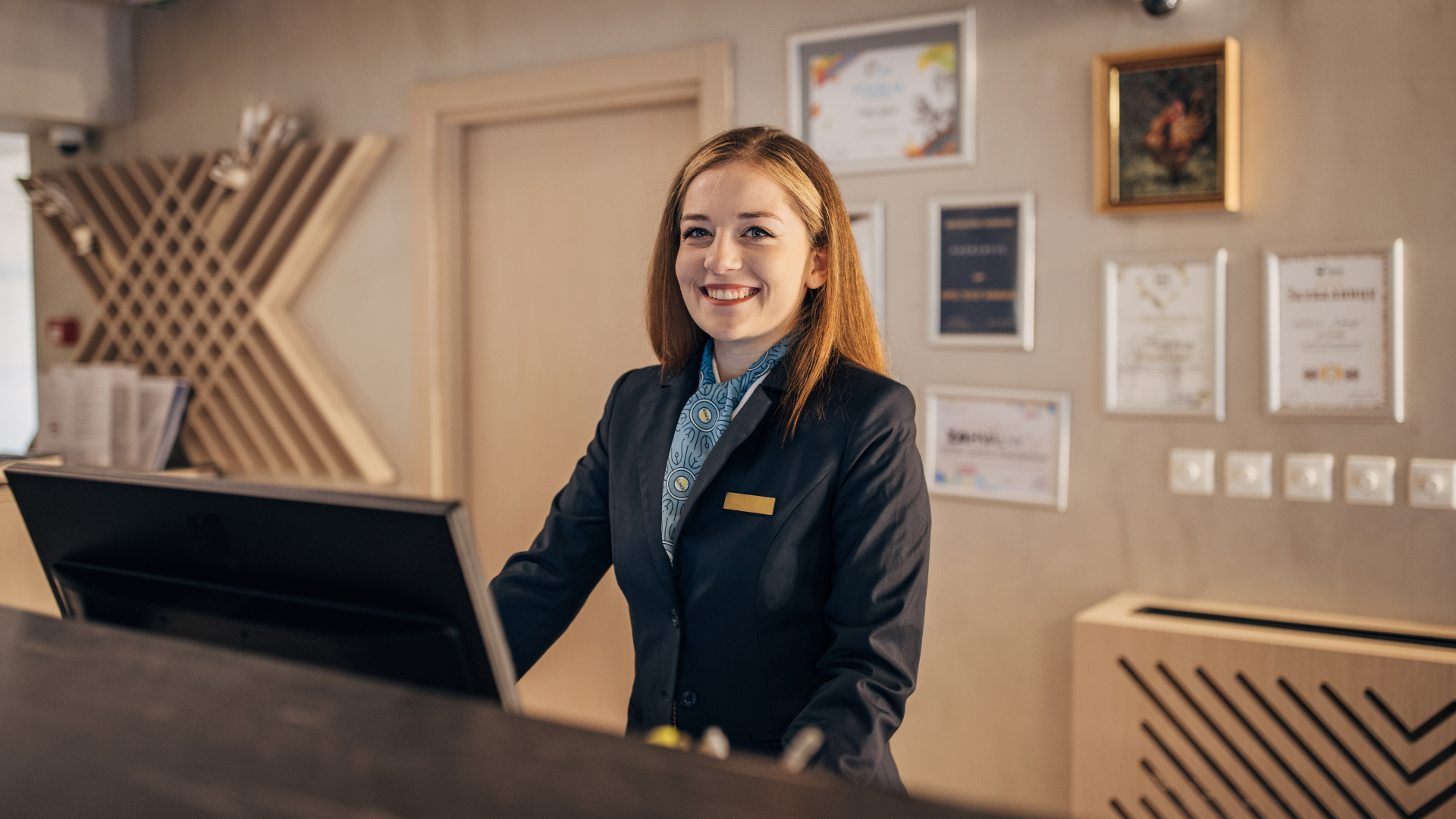 A smiling receptionist in a navy blazer stands behind a front desk in a modern lobby, ready to assist with your hotel booking. Framed certificates and awards are displayed on the wall behind her.