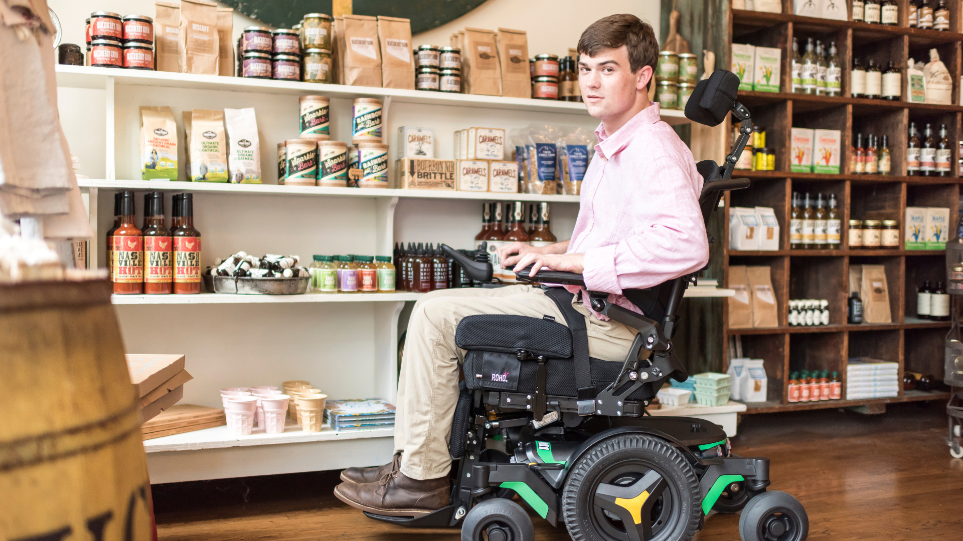A young man in a pink shirt uses a powered wheelchair with built-in pressure relief features inside a store, surrounded by shelves stocked with food items, jars, and bottles. He looks toward the camera with a neutral expression.