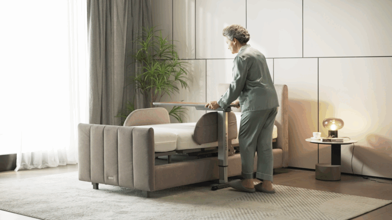 An elderly woman in green pajamas stands beside an adjustable bed with quality bedding, holding a movable table. The modern, well-lit room features a potted plant, light gray curtains, and a small side table with a mug and lamp.