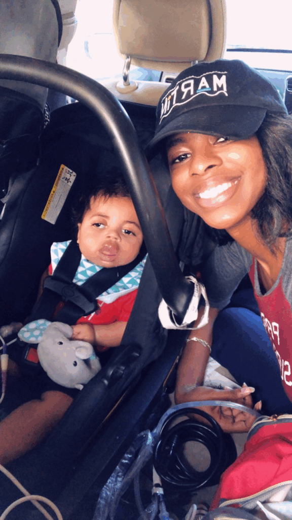 A smiling woman wearing a black hat sits next to a baby in a car seat. The baby, secured with a harness and holding a gray stuffed animal, is content as daylight streams through the car windows.