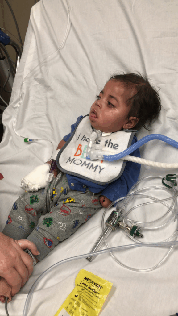 A young child lies in a hospital bed with an oxygen tube and IV, wearing a bib that says I have the best mommy. Medical equipment and a yellow packet are visible nearby as an adult offers comforting support.