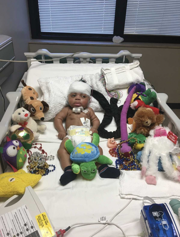 A baby lies in a hospital bed with medical bandages and equipment, surrounded by colorful stuffed animals, beads, and sensory toys. The room has a window with closed blinds in the background.