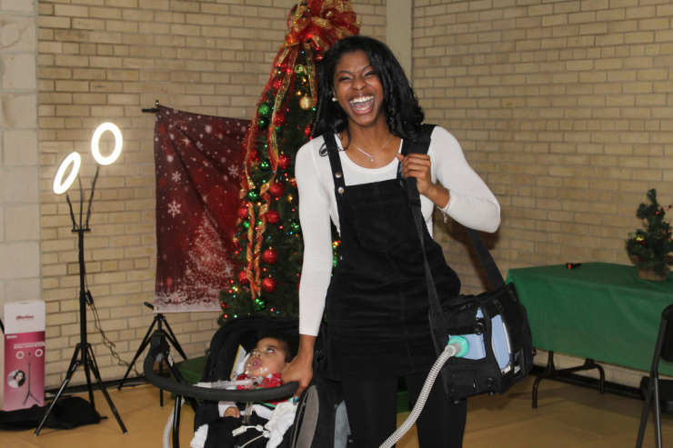 A woman smiles brightly while pushing a young child in a stroller. Behind them is a decorated Christmas tree with festive decorations, a red bow, and a ring light. The indoor setting feels warm and joyful.