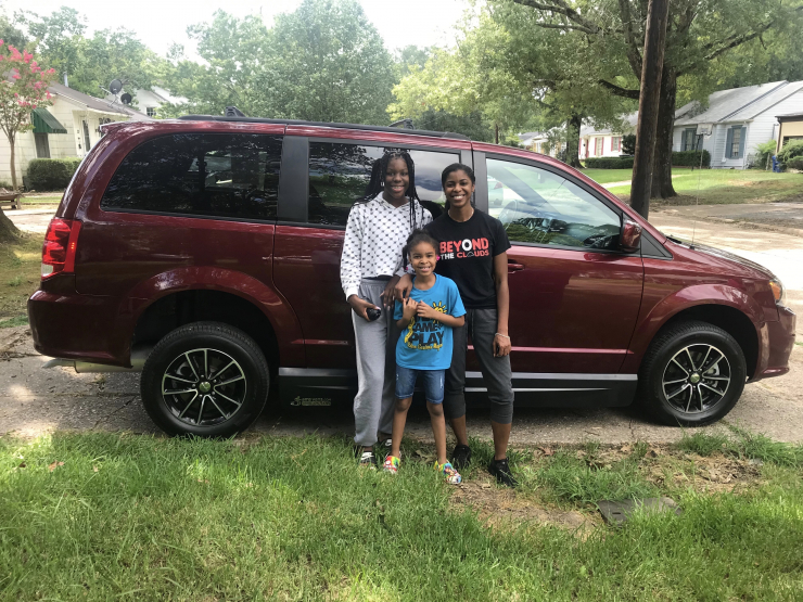 Three people, two adults and one child, stand smiling in front of a parked maroon accessible minivan on a quiet residential street lined with trees and houses.