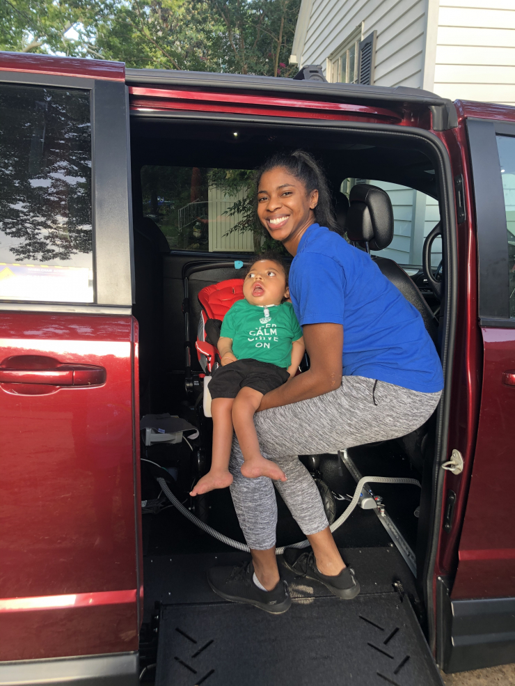 A smiling woman in a blue shirt sits on the edge of an open red van next to a young child in a green shirt, safely seated in a car seat. Trees and a house are visible in the background.