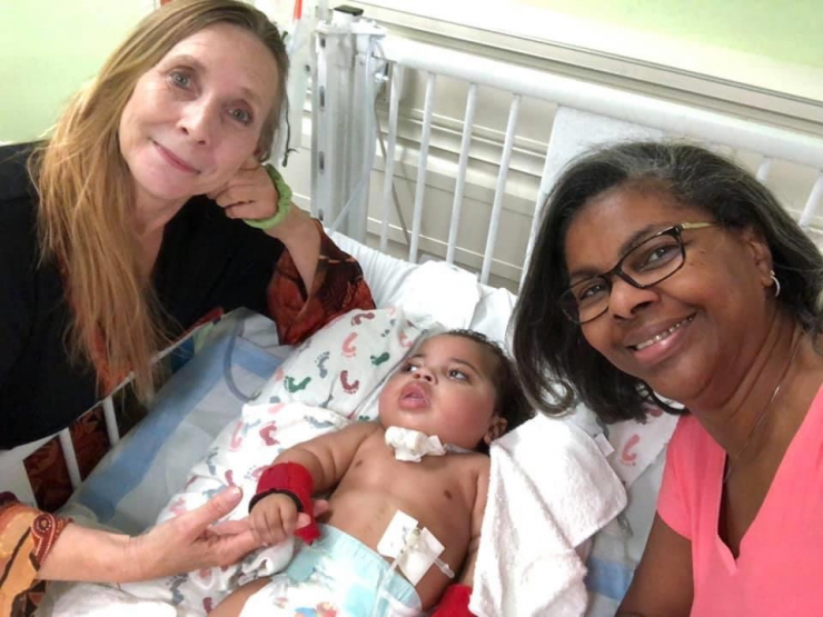 Two women smile at the camera while sitting beside a baby in a hospital crib. The baby, with medical equipment attached, lies on a blanket as one woman gently holds the babys hand.