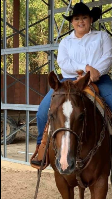 A person wearing a black cowboy hat, white shirt, and jeans sits smiling on a brown horse with a white blaze inside an open metal building. Trees are visible in the background, capturing a moment from Analieses Story.