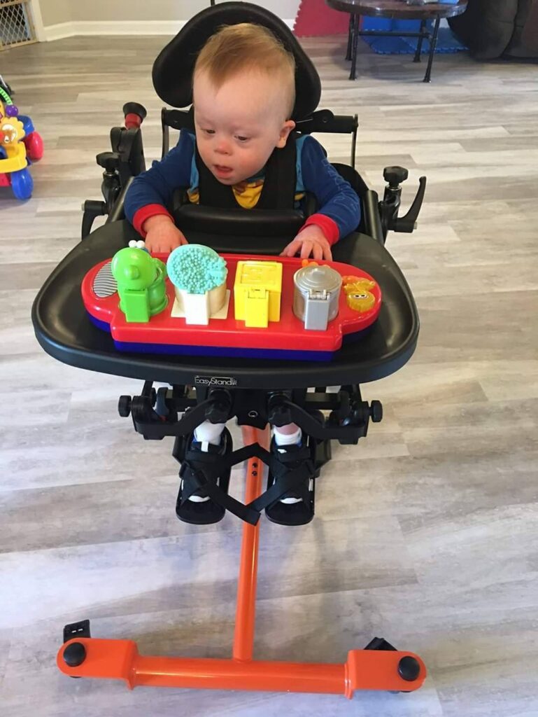 A young child enjoys colorful toys on a tray while using a specialized Levis standing mobility device with supportive straps, in a room with light wood flooring and toys in the background, creating a joyful story of play.