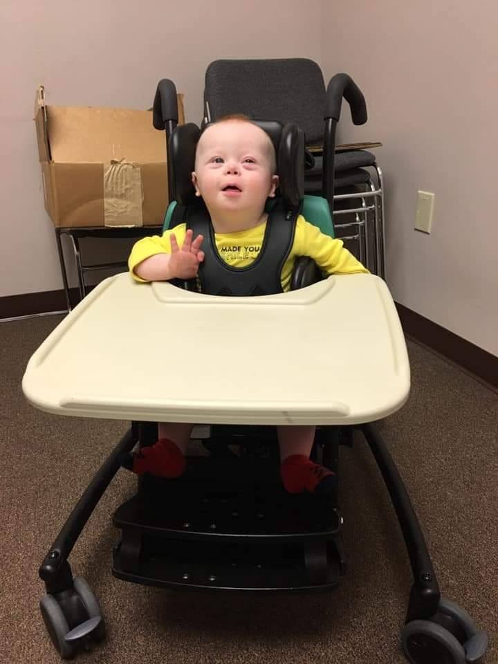 A young child with Down syndrome sits in a wheelchair behind a tray, wearing a yellow Levis outfit and smiling while waving. The background tells the story of everyday life, with a cardboard box and stacked chairs against beige walls.