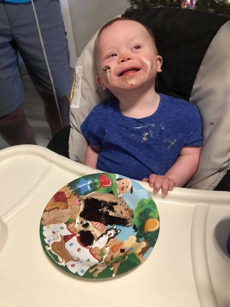 A young child with cake smeared on their face and Levis shirt smiles joyfully while sitting in a high chair. A colorful plate with a piece of chocolate and vanilla cake sits on the tray, ready for the next bite in this sweet story.