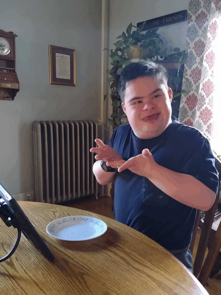 A young man with Down syndrome sits at a wooden table, smiling and signing with his hands. Calebs Story unfolds as he enjoys a decorative plate in front of him, with a tablet nearby in a warmly lit room filled with plants and wall decor.