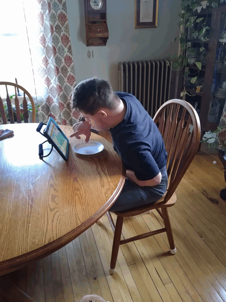 A boy sits at a wooden dining table, eating from a bowl while watching Calebs Story on a tablet propped up on a stand. The room features floral curtains, a wall clock, framed art, a radiator, and houseplants.