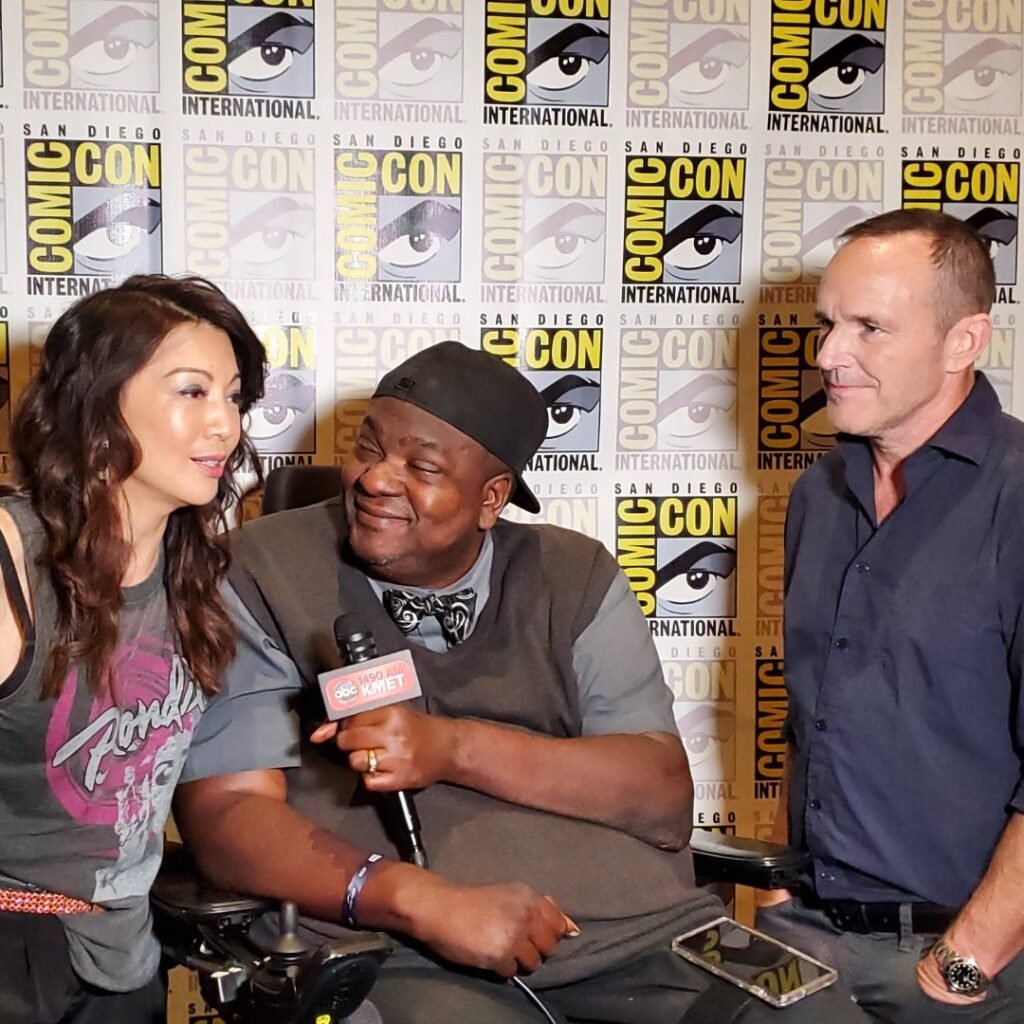 Three people are smiling and talking during an interview at San Diego Comic-Con, discussing Pauls Story. They stand in front of a Comic-Con International backdrop, with one person holding a microphone.