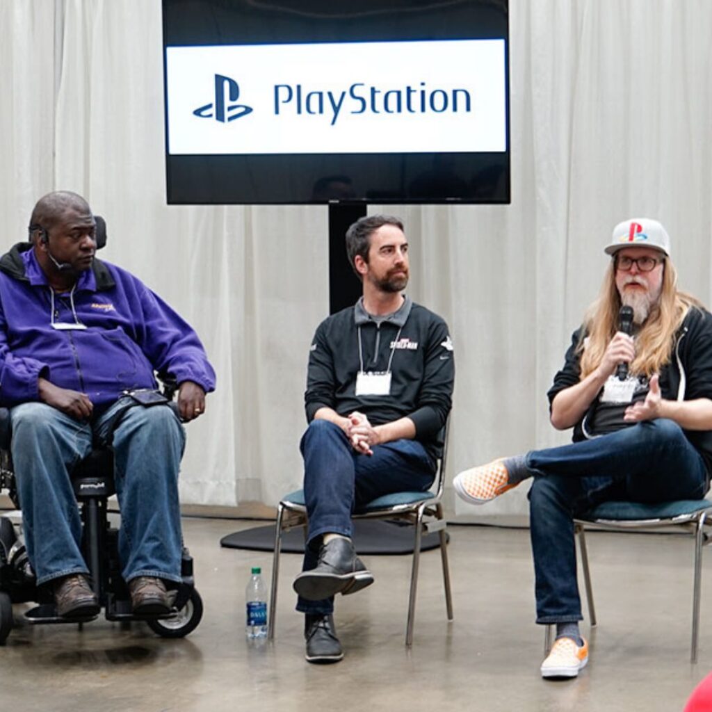 Three men sit on chairs in front of a PlayStation sign; one uses a wheelchair, another holds a microphone and gestures while speaking about Pauls Story, and the third listens attentively. A water bottle rests on the floor beside them.