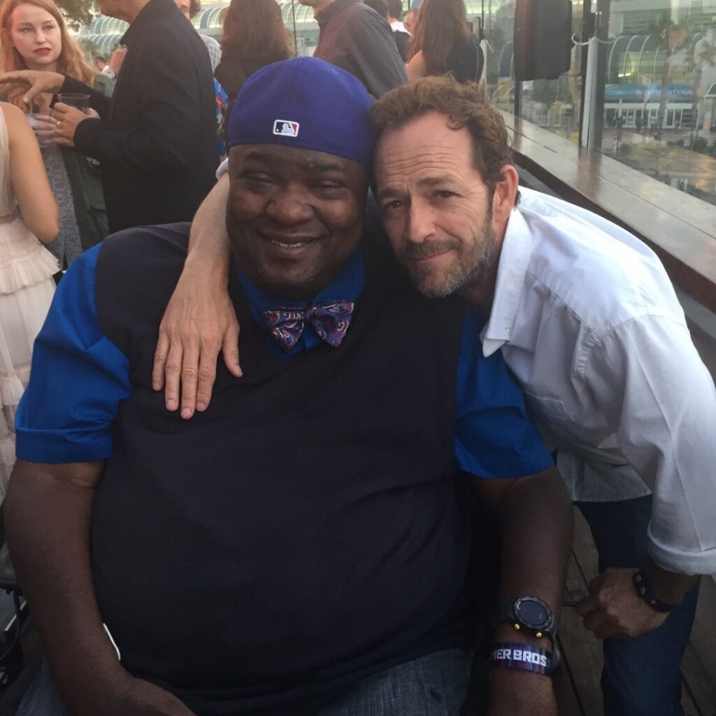 Two men pose together at an outdoor event—one in a blue shirt, bowtie, and baseball cap, the other in a white shirt with his arm around him. Pauls Story unfolds against a lively cityscape backdrop with people mingling nearby.