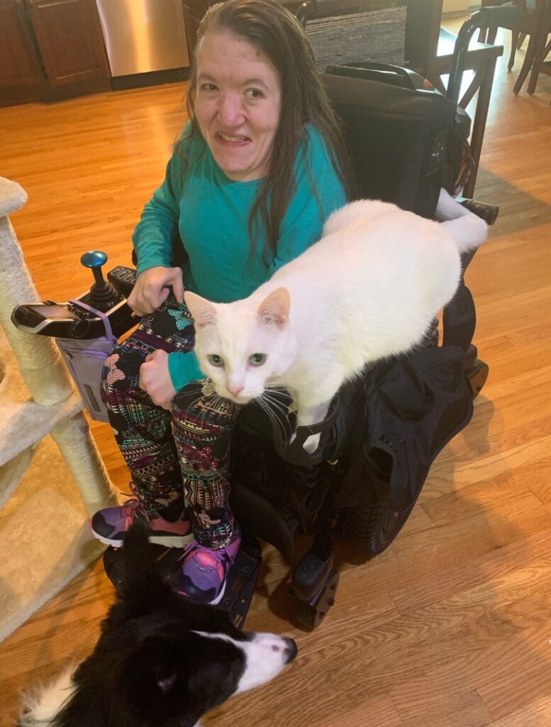 Rebeccas Story: A woman in a wheelchair smiles at the camera. A large white cat rests on her lap, while a black and white dog lies nearby on cozy wooden floors.