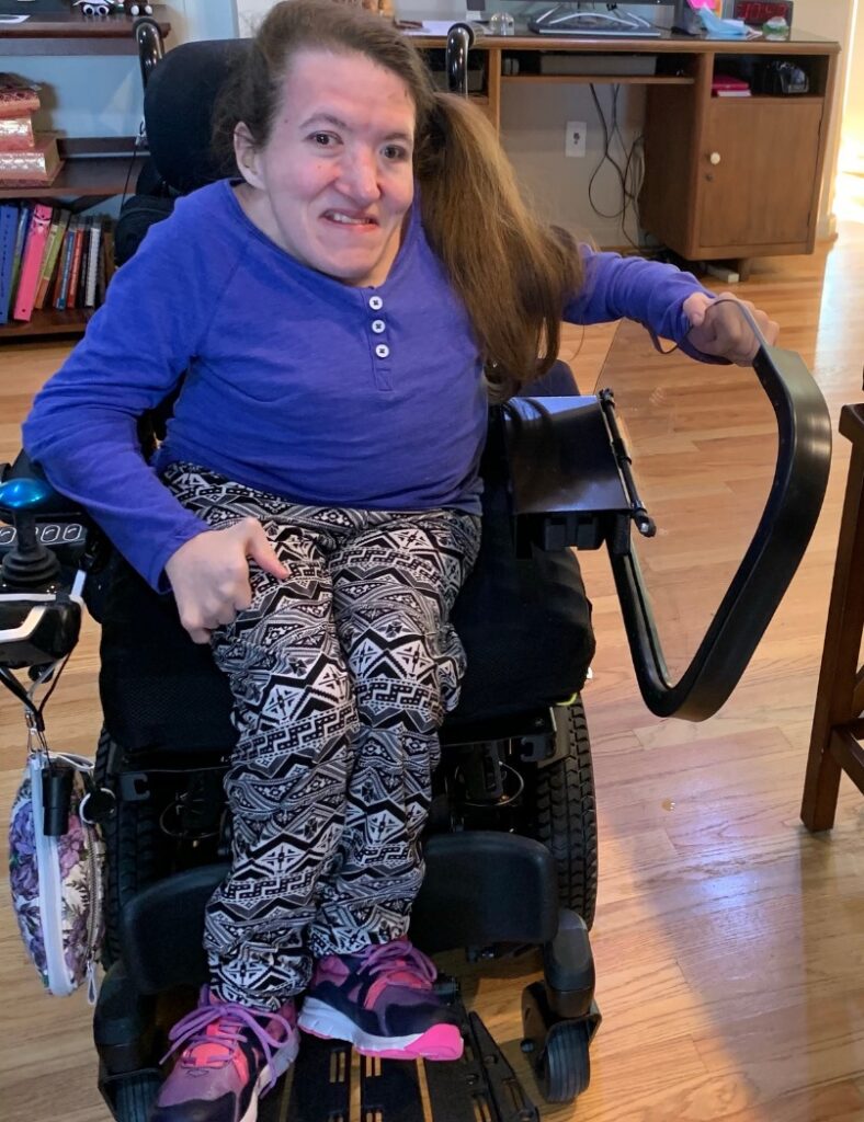 Rebecca’s Story: A woman in a patterned shirt and pants sits in a motorized wheelchair indoors, smiling at the camera. She has long brown hair, wears pink sneakers, and holds the armrest. Wooden floors and bookshelves are in the background.