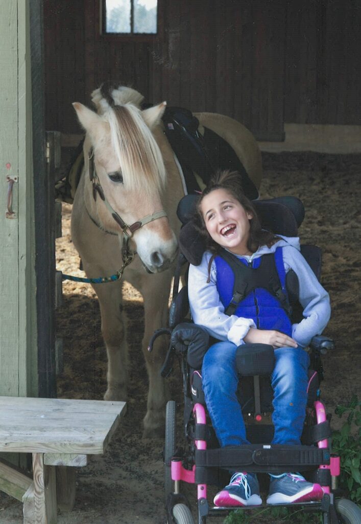 Lexis Story: A smiling girl in a wheelchair sits in a barn doorway, wearing a blue vest. Behind her stands a light brown horse with a cream-colored mane, looking out from a stall.