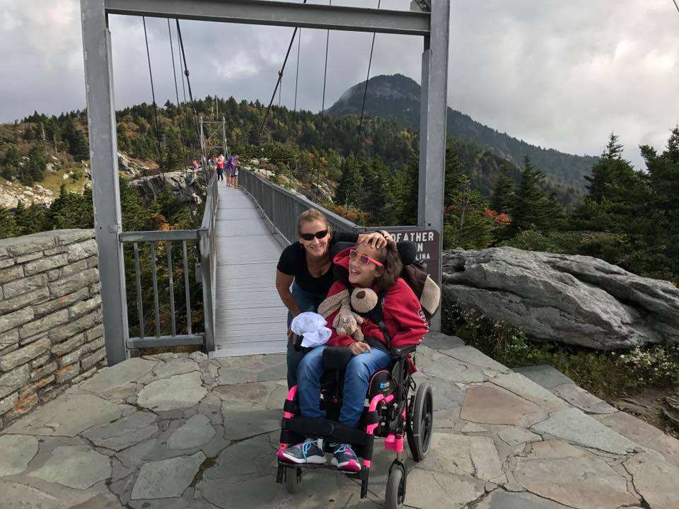 A woman stands smiling behind a girl in a wheelchair, who is holding a stuffed animal and smiling, at the entrance to a suspension bridge with mountains and trees in the background. This moment captures Lexis Story of joy and adventure.