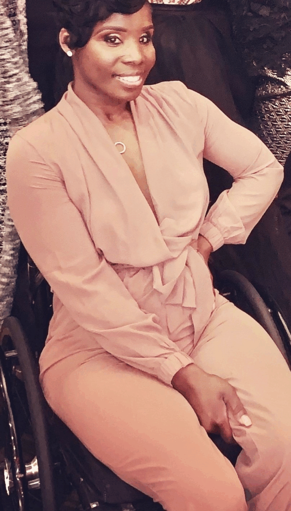 A woman wearing a light pink, long-sleeved jumpsuit sits in a wheelchair, smiling at the camera with one hand on her hip—capturing Nakias Story.