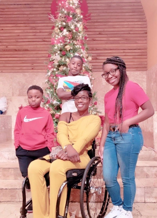 Four people pose in front of a decorated Christmas tree. A woman in a yellow outfit sits in a wheelchair, while two boys and a girl stand around her, all smiling. The festive scene captures the warmth and togetherness of Nakias Story.