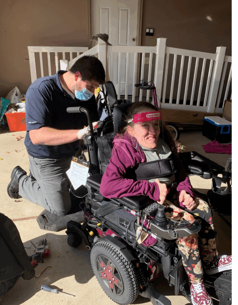 A man wearing a face mask kneels beside a smiling young woman in a motorized wheelchair, adjusting equipment attached to her chair. They are outdoors near a porch with white railings, sharing a moment from Rebeccas Story.