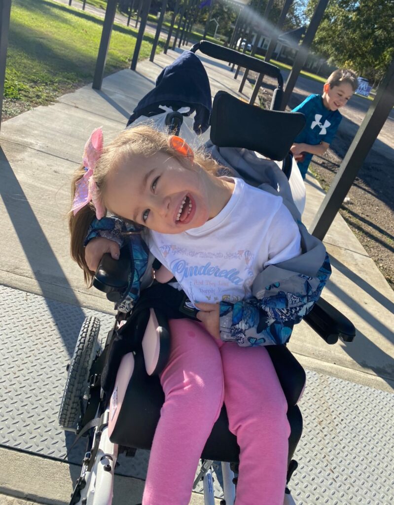 A young girl named Avery Joy with a pink bow smiles in her wheelchair outdoors on a sunny day. She wears a white Cinderella shirt and pink pants. A smiling boy stands in the background, adding to the cheerful story.