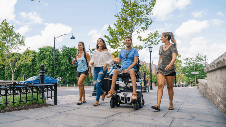 Four young adults walk and laugh together on a sunny sidewalk. One person is a wheelchair user, and everyone appears cheerful, enjoying their time outdoors. Trees and a street are visible in the background.