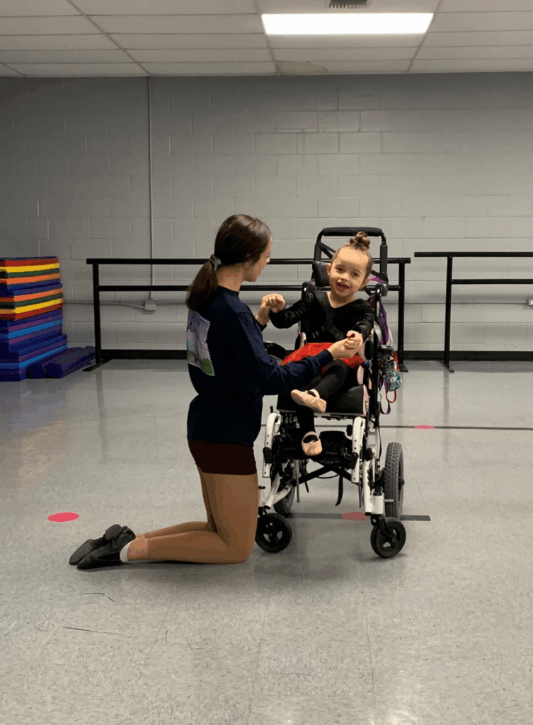 A young woman kneels on the floor, holding hands with a smiling girl named Avery Joy in a wheelchair inside a dance studio. Ballet barres and mats set the scene for their inspiring story.