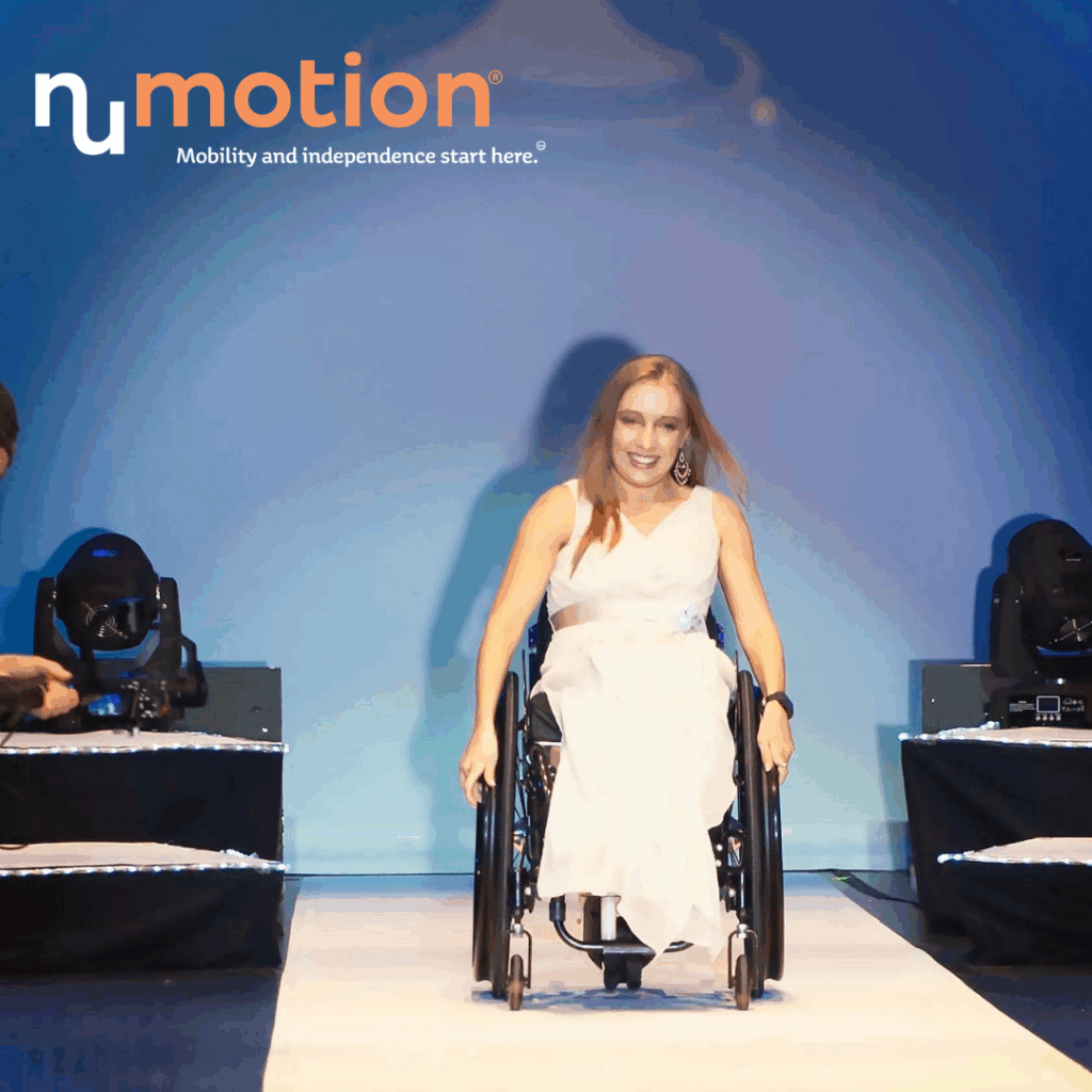 A woman in a white dress confidently rolls down a runway in a wheelchair under stage lights, with the NuMotion logo and Kyanns Story: Mobility and independence start here above her.