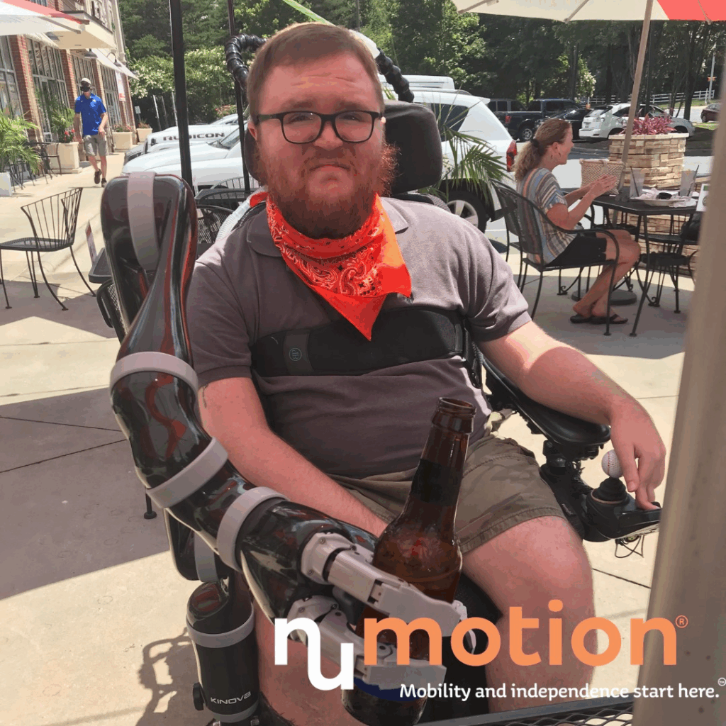 Seans Story: A man in a wheelchair with a robotic arm attachment holds a beer bottle outdoors at a cafe. He wears glasses, a red bandana, and a gray shirt. NuMotion’s logo and slogan appear in the lower right corner.