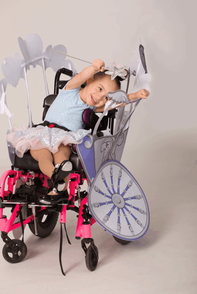 Avery Joy, a young girl in a sparkling blue dress and star headband, smiles and poses as her wheelchair—decorated as a whimsical, fairy-tale carriage with purple and silver accents—brings her story to life.