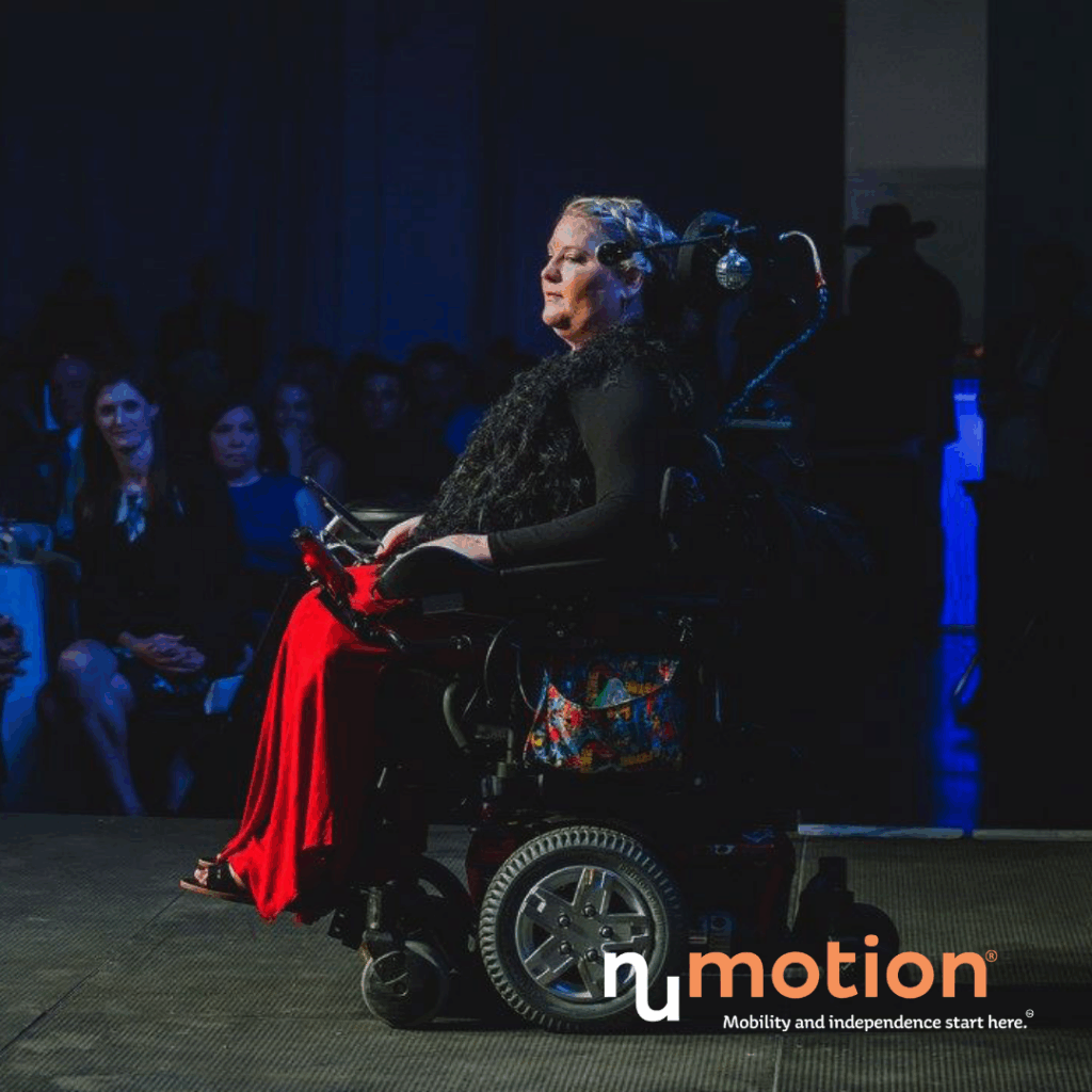 A woman in a power wheelchair wearing a black top and red skirt moves confidently down a runway, watched by an audience. Soft blue lighting sets the scene. The NuMotion logo and slogan appear in the lower right corner.