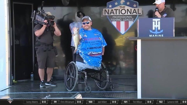 A man in a wheelchair proudly holds up a large fish on stage at a National Professional Fishing League event, sharing Hunters Story as a cameraman films and an announcer speaks at the podium.