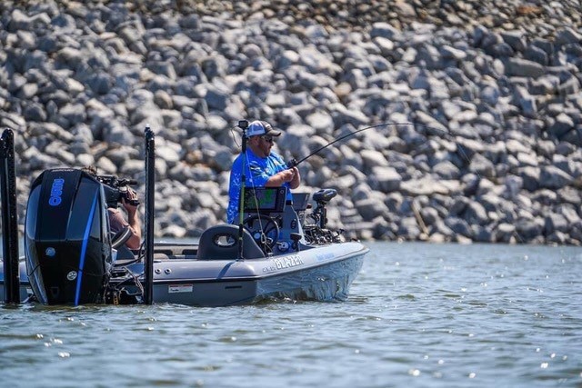 A person in a blue shirt and cap is fishing from a motorboat on calm water, with a rocky shoreline in the background, capturing the essence of a true Hunters Story.