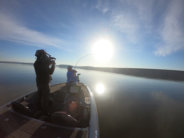 A person fishes from a boat on calm water at sunrise or sunset, while another films them with a camera. The sky is mostly clear, sunlight reflects on the water’s surface—capturing a tranquil moment in this Hunters Story.