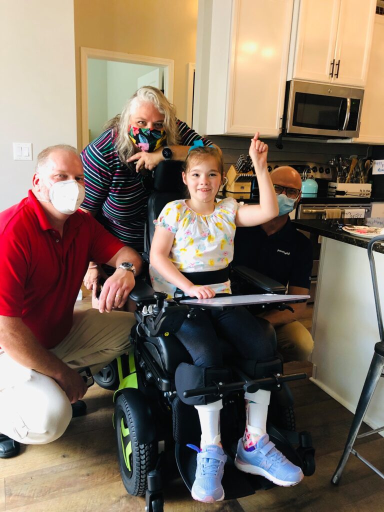 A young girl in a wheelchair smiles and points upward, surrounded by three adults wearing face masks in a kitchen. This joyful moment captures Zoeys Story, set against bright lighting and modern appliances.