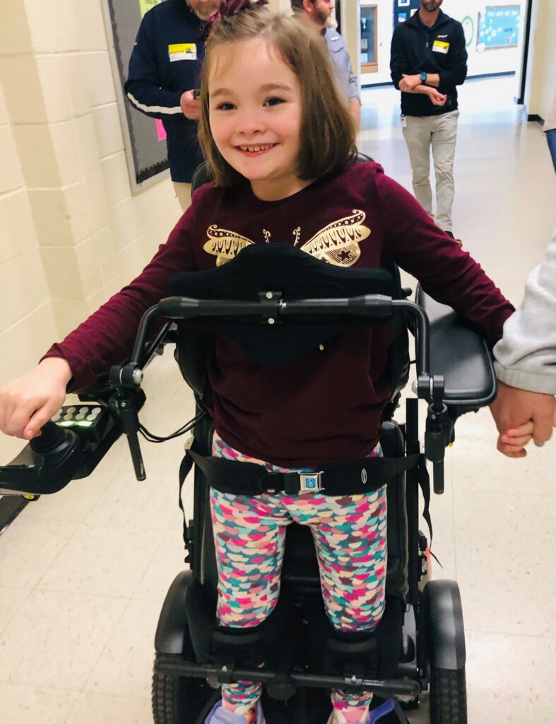 Zoeys Story: A young girl with brown hair smiles as she uses a motorized wheelchair. She wears a maroon shirt with gold wings and colorful leggings, holding hands with an adult in a hallway.