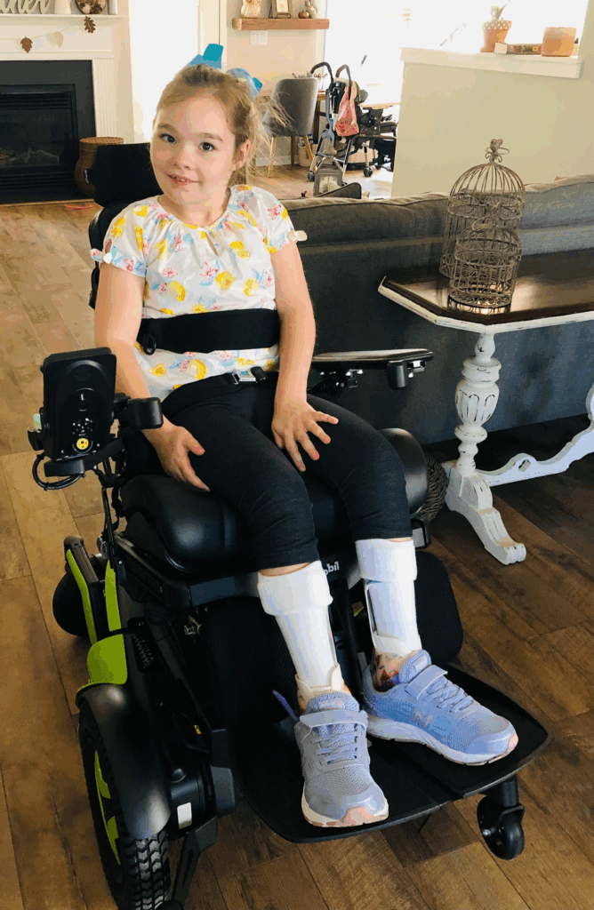 A young girl with brown hair in a ponytail sits in a motorized wheelchair, smiling. Part of Zoeys Story, she wears a colorful shirt, dark pants, orthopedic leg braces, and purple sneakers on a wooden floor near a table and birdcage.