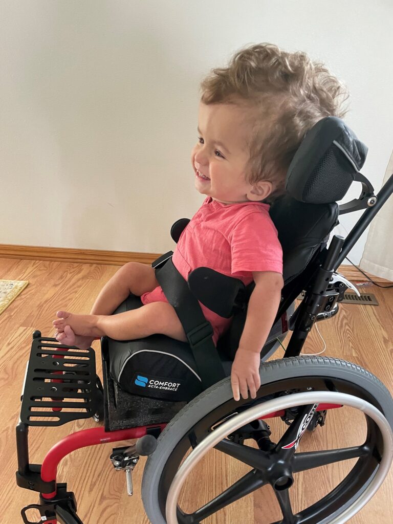 A young child with curly hair, wearing a pink shirt, smiles while sitting in a black and red wheelchair on a wooden floor indoors as part of Henrys Story.