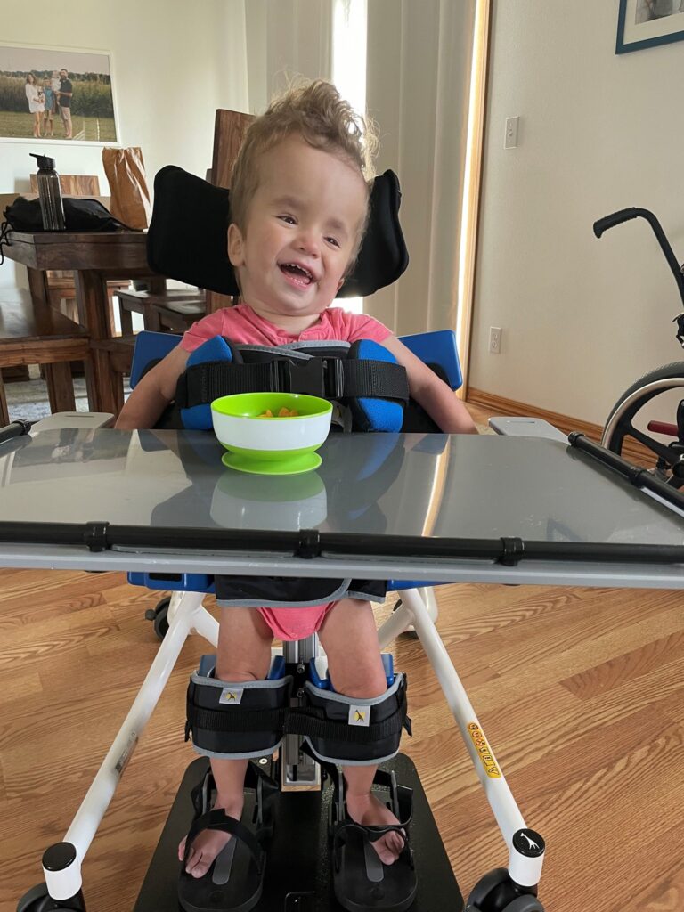 A young child with curly hair sits in a supportive wheelchair at a tray table, smiling and wearing a pink shirt. A green bowl is on the tray. Henrys Story unfolds in a room with wooden floors and furniture in the background.