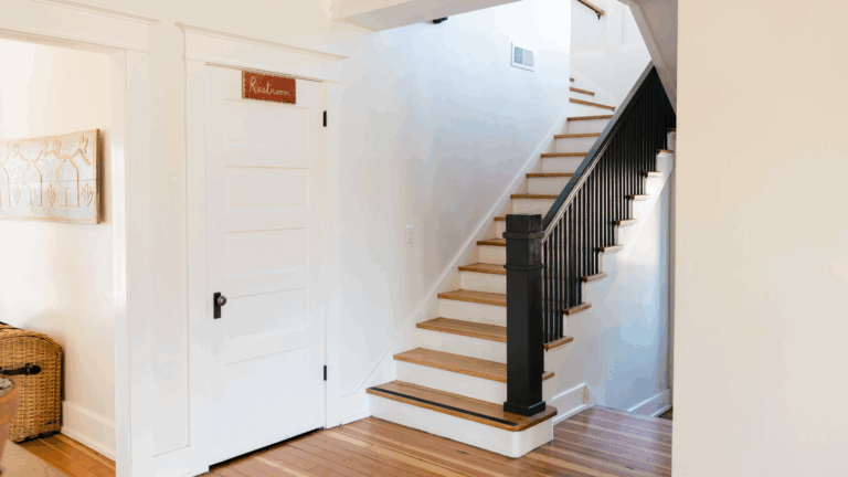 A bright hallway with wooden floors, a white door on the left, and a staircase featuring wooden steps and black railings designed for stairs safety. Light fills the space, creating a clean and welcoming atmosphere.