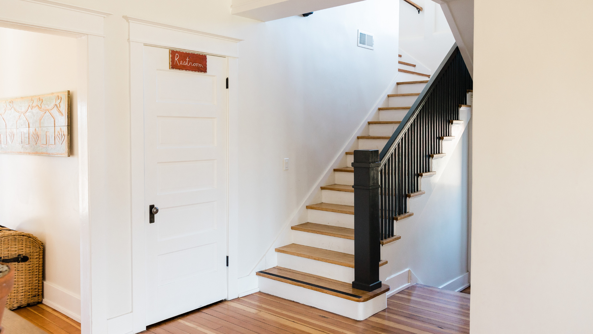A bright hallway with wooden floors, a white door on the left, and a staircase featuring wooden steps and black railings designed for stairs safety. Light fills the space, creating a clean and welcoming atmosphere.