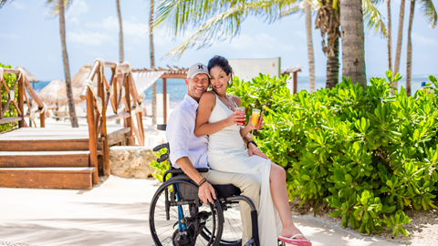 Kriss Story: A man using a wheelchair and a woman in a white dress smile and hold drinks while sitting together on a sunny tropical beach with palm trees and ocean in the background.