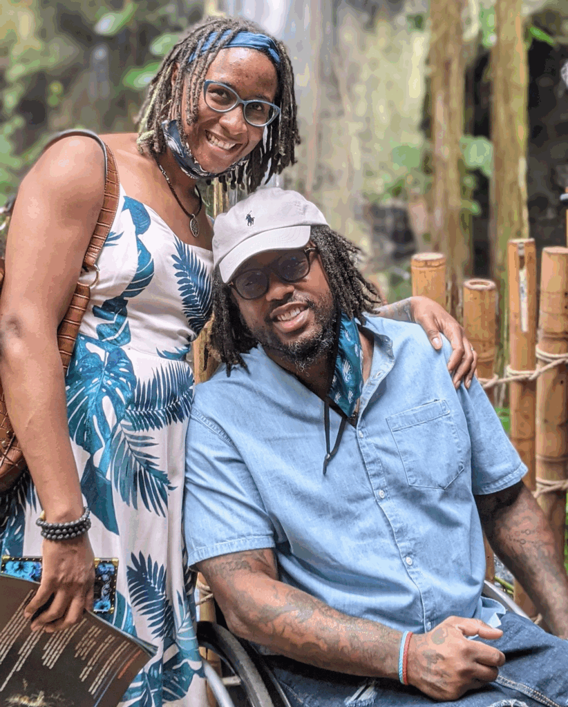 A smiling woman in a tropical dress stands beside a smiling man in a wheelchair wearing a light blue shirt and white cap, surrounded by lush greenery and bamboo—capturing a joyful moment from Lionels Story.